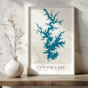 Könnte beinhalten: Ein gerahmter vertikaler Wandkunstdruck mit einer blaugrünen Karte eines Sees auf cremefarbenem Hintergrund. Der Text am unteren Rand lautet CUSTOM LAKE, NORTH CAROLINA, 35.486 N, 80.923 W. Der Druck befindet sich in einem einfachen weißen Rahmen.