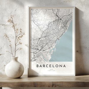 May include: A framed vertical wall art print featuring a detailed black and white street map of Barcelona, Spain. The map displays intricate road networks against a light background, with a pale blue sea area. Text reads BARCELONA SPAIN 41.387°N | 2.168°E.