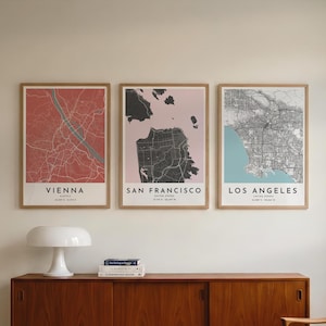 Op de afbeelding: Drie ingelijste wandposters met gestileerde stadsplattegronden van Wenen, San Francisco en Los Angeles. Elke print toont de stadsnaam, het land en de geografische coördinaten in zwarte tekst onderaan op een gekleurde achtergrond.