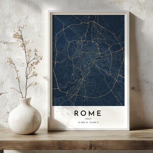 Puede incluir: Una lámina de arte enmarcada que muestra un mapa de Roma, Italia, en azul oscuro con líneas de calles doradas. El texto en la parte inferior dice ROME, ITALY, y las coordenadas 41.903° N - 12.496° E. La lámina está en un marco blanco.