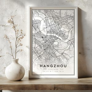 May include: A framed vertical wall art print featuring a black and white street map of Hangzhou, Zhejiang, with coordinates 30.27 N | 120.15 E printed at the bottom. The map displays a detailed network of roads and a river flowing through the city.