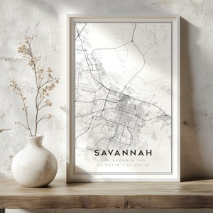 Op de afbeelding: Een ingelijste wandkunstprint met een zwart-wit stadsplattegrond van Savannah, Georgia. De print bevat de stadsnaam, de staat en de geografische coördinaten 32.081 N | 81.091 W in een strak, schreefloos lettertype onderaan in het midden.