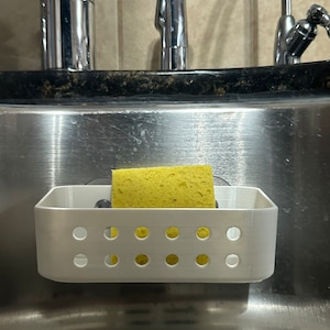 May include: White plastic dish rack with a yellow sponge inside, mounted to a stainless steel kitchen sink.