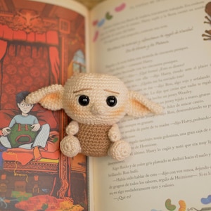 Może przedstawiać: Szydełkowa lalka postaci z dużymi uszami i czarnymi oczami, siedząca na otwartej książce. Lalka jest beżowa i brązowa, o szczegółowym wzorze. Książka ma kolorowe ilustracje i tekst.