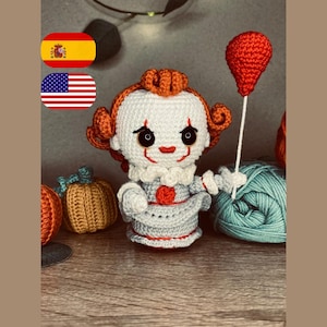 Peut inclure: Poupée clown au crochet avec des cheveux orange, un col blanc à volants et une tenue grise. La poupée tient un ballon rouge sur une ficelle blanche. Des citrouilles crochetées et une pelote de fil turquoise sont également visibles.