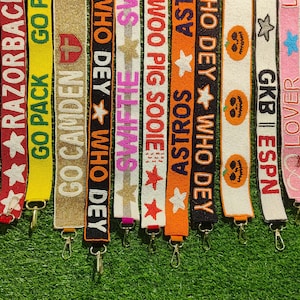 May include: A collection of beaded bag straps in various colors, each featuring text and designs. The straps display phrases like "GO HOGS," "RAZORBACKS," and "ASTROS," along with stars, pumpkins, and other decorative elements. Each strap has a metal clasp.
