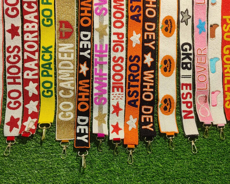 May include: A collection of beaded bag straps in various colors, each featuring text and designs. The straps display phrases like "GO HOGS," "RAZORBACKS," and "ASTROS," along with stars, pumpkins, and other decorative elements. Each strap has a metal clasp.
