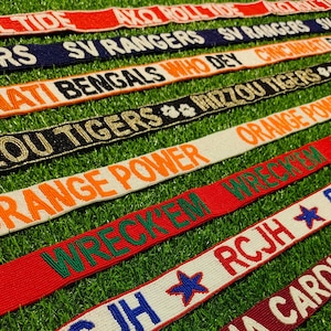 May include: Close-up of several beaded ribbons with team names and logos. The ribbons are in various colors, including red, orange, black, and white, with text like "ROLL TIDE" and "ORANGE POWER". They are arranged on a green surface.