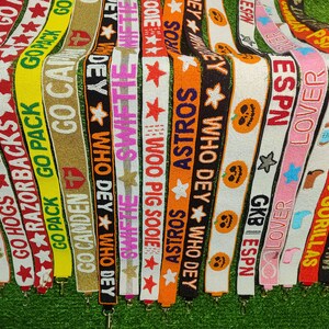 May include: A collection of beaded bag straps in various colors, each featuring text and star designs. The straps display phrases like "GO HOGS," "ASTROS," and "ESPN," with a variety of colors including red, orange, and pink.