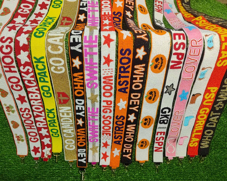 May include: A collection of beaded bag straps in various colors, each featuring text and star designs. The straps display phrases like "GO HOGS," "ASTROS," and "ESPN," with a variety of colors including red, orange, and pink.