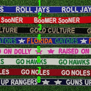 Puede incluir: Una colección de cintas con cuentas con varias frases y nombres de equipos. Las cintas son de diferentes colores, incluyendo azul, rojo, negro y blanco, con textos como "ROLL JAYS" y "GO HAWKS".