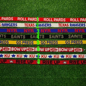 Puede incluir: Una colección de ocho cordones de tela con varios nombres de equipos y frases impresas. Los cordones son de diferentes colores, incluyendo rojo, blanco, azul, negro y amarillo. El texto incluye "ROLL PARDS", "TEXAS RANGERS", "NYR", "SAINTS", "GO PREDS", "BOW UP", "KU" y "RISE UP".