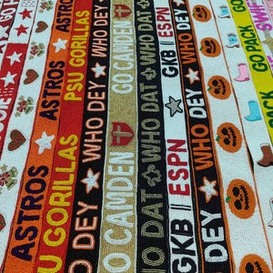 May include: A collection of beaded ribbons in various colors, featuring text and designs. The ribbons display phrases like "GO HOGS," "ASTROS," and "WHO DEY," along with stars, hearts, and pumpkin motifs. The overall theme is sports and seasonal.