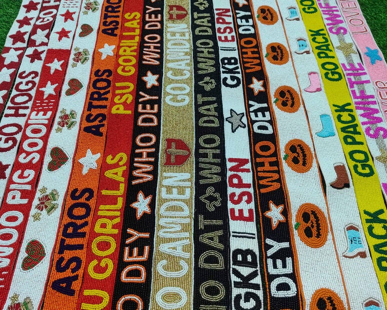 May include: A collection of beaded ribbons in various colors, featuring text and designs. The ribbons display phrases like "GO HOGS," "ASTROS," and "WHO DEY," along with stars, hearts, and pumpkin motifs. The overall theme is sports and seasonal.