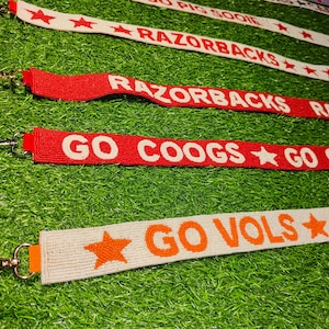 Op de afbeelding: Close-up van diverse lanyards met teamnamen en sterren. De lanyards zijn rood, wit en oranje, met teksten als "RAZORBACKS", "GO COOGS" en "GO VOLS". Elke lanyard heeft een metalen sluiting.