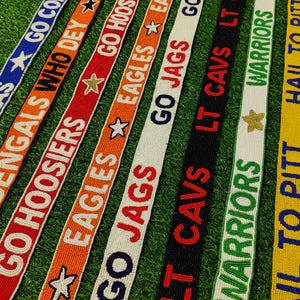 May include: A collection of beaded team spirit straps in various colors, including blue, orange, red, black, white, and yellow. Each strap features team names and phrases like "GO COBRAS" and "HAIL TO PITT" in bold lettering, with star accents.