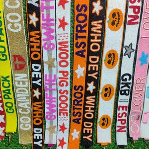 May include: A collection of beaded lanyards in various colors, featuring text and designs. The lanyards display phrases like "GO PACK", "ASTROS", and "WHO DEY", along with star and pumpkin motifs. Each lanyard has a metal clasp.