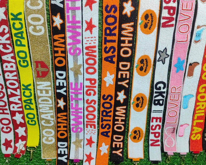 May include: A collection of beaded lanyards in various colors, featuring text and designs. The lanyards display phrases like "GO PACK", "ASTROS", and "WHO DEY", along with star and pumpkin motifs. Each lanyard has a metal clasp.