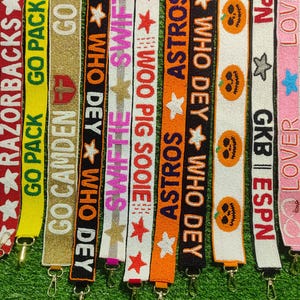 May include: A collection of beaded lanyards in various colors, each featuring text and designs. The lanyards display phrases like "GO HOGS," "RAZORBACKS," and "ASTROS," along with star and pumpkin motifs. Each lanyard has a metal clasp.