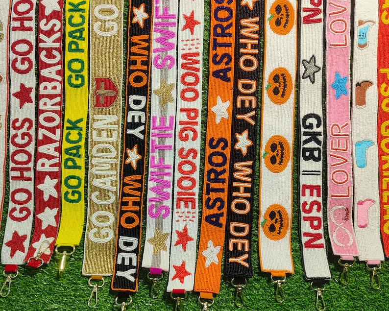 May include: A collection of beaded lanyards in various colors, each featuring text and designs. The lanyards display phrases like "GO HOGS," "RAZORBACKS," and "ASTROS," along with star and pumpkin motifs. Each lanyard has a metal clasp.