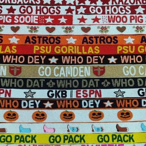 May include: A collection of beaded ribbons with various text and designs. The ribbons feature phrases like "GO HOGS", "ASTROS", and "WHO DEY", along with star and pumpkin motifs. The color palette includes red, orange, black, and yellow.