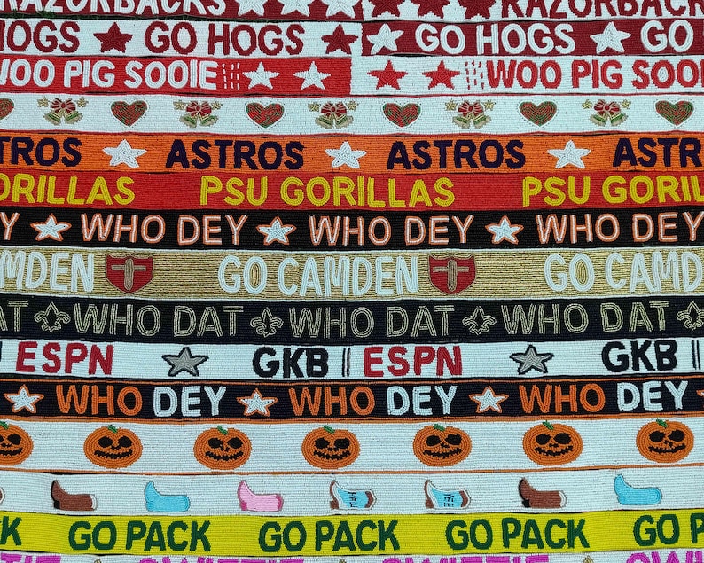 May include: A collection of beaded ribbons with various text and designs. The ribbons feature phrases like "GO HOGS", "ASTROS", and "WHO DEY", along with star and pumpkin motifs. The color palette includes red, orange, black, and yellow.