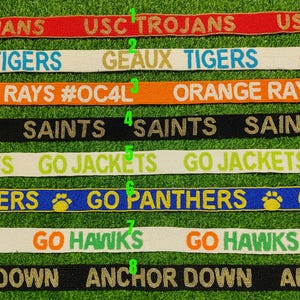 May include: A collection of beaded ribbons with team names and phrases. The ribbons are in various colors, including red, white, orange, black, blue, and green. Phrases include "USC Trojans", "Geaux Tigers", "Orange Rays", "Saints", "Go Jackets", "Go Panthers", "Go Hawks", and "Anchor Down".