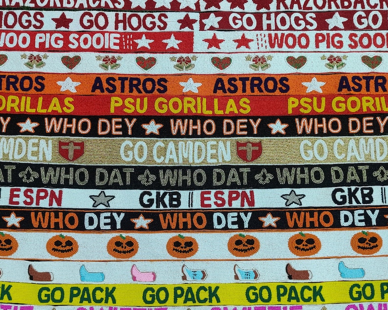 May include: A collection of beaded ribbons with various designs and text. The ribbons feature phrases like "GO HOGS," "ASTROS," and "WHO DEY," along with star and heart motifs. The color palette includes red, orange, black, and white.