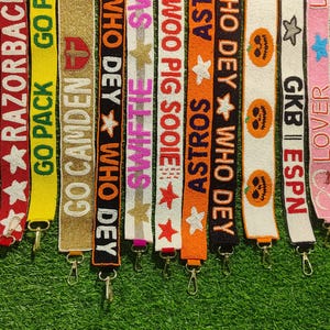 May include: A collection of beaded bag straps in various colors, each featuring text and designs. The straps include phrases like "GO HOGS," "RAZORBACKS," and "ASTROS," along with stars, pumpkins, and other decorative elements. Each strap has a metal clasp.