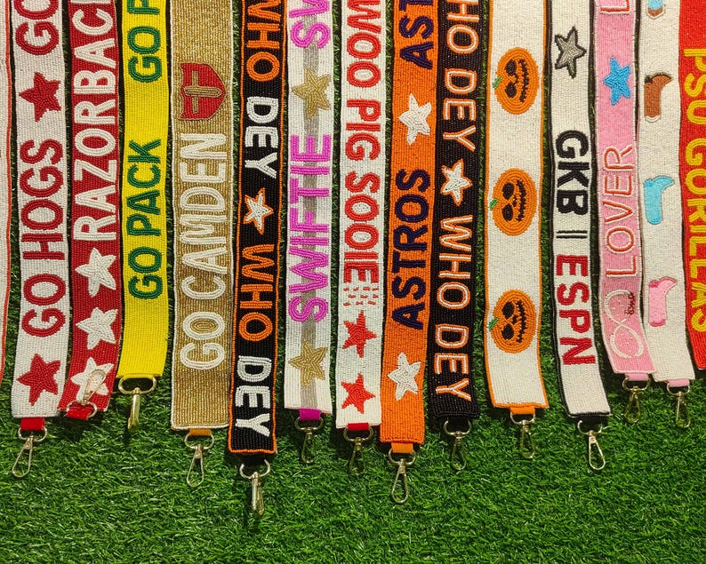 May include: A collection of beaded bag straps in various colors, each featuring text and designs. The straps include phrases like "GO HOGS," "RAZORBACKS," and "ASTROS," along with stars, pumpkins, and other decorative elements. Each strap has a metal clasp.
