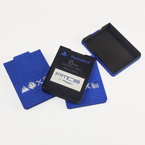 May include: A black 8MB PlayStation 2 memory card with the text "PlayStation 2" and "8MB Memory Card MagicGate" is displayed. The card is next to two blue cases with the text "AOX" in white.