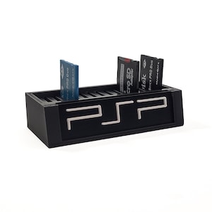 以下が含まれることがあります： 黒いPSPメモリーカードホルダーに3枚のメモリーカードが挿入されています。ホルダーには白いPSPロゴがあります。メモリーカードには「Stick Pro Duo」と「Micro SD Adaptor」と記載されています。