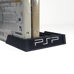 Puede incluir: Un soporte negro para juegos de PSP con el logotipo de PSP en blanco. Hay dos cajas de juegos en el soporte. Las cajas de juegos son blancas y beige. El soporte para juegos está sobre una superficie blanca.