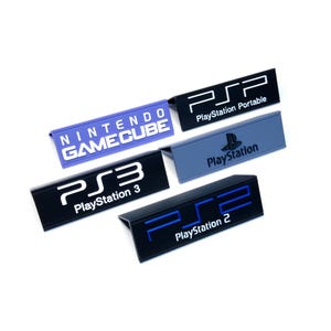 May include: A collection of black, blue, and purple game console nameplates. The nameplates include "PSP PlayStation Portable", "PlayStation 2", "PlayStation 3", "PlayStation", and "Nintendo GameCube". The text is white or blue.