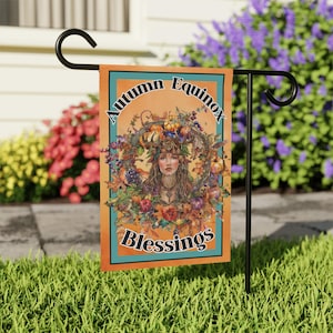 Puede incluir: Una bandera de jardín con fondo naranja y borde turquesa. La bandera presenta a una mujer con cabello largo y castaño que lleva una corona de hojas de otoño, frutas y verduras. El texto "Autumn Equinox Blessings" está impreso en la bandera.