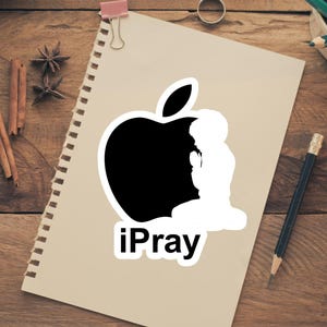 May include: A sticker featuring a black apple silhouette with a white praying figure inside. The text "iPray" is below the apple. The sticker is on a notepad, with pencils, cinnamon sticks, and other items on a wooden surface.