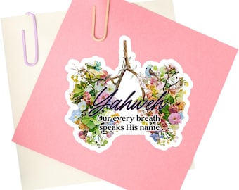 May include: A sticker with the word "Yahweh" in black script, surrounded by a floral design with a small bird. The sticker is on a pink paper, held by a pink paper clip, with a white paper underneath, held by a purple paper clip.