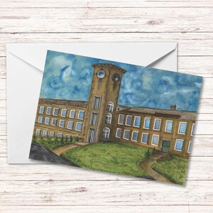 Puede incluir: Pintura a la acuarela de un edificio grande y marrón con una torre de reloj, sobre un cielo azul con nubes. El edificio tiene muchas ventanas y un césped verde en frente. La obra de arte está en una tarjeta dentro de un sobre blanco.