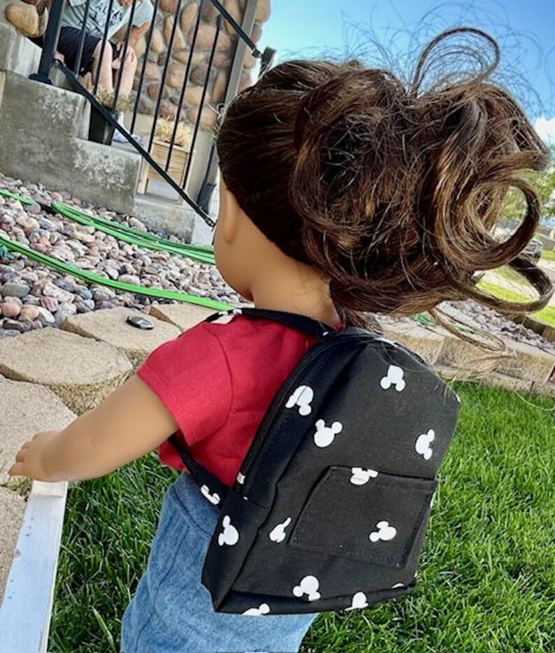 May include: A black backpack with white Mickey Mouse silhouettes all over it. The backpack has a small pocket on the front.