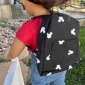 May include: A black backpack with white Mickey Mouse heads printed all over it. The backpack has a small pocket on the front. The backpack is being worn by a doll with brown hair and a red shirt.
