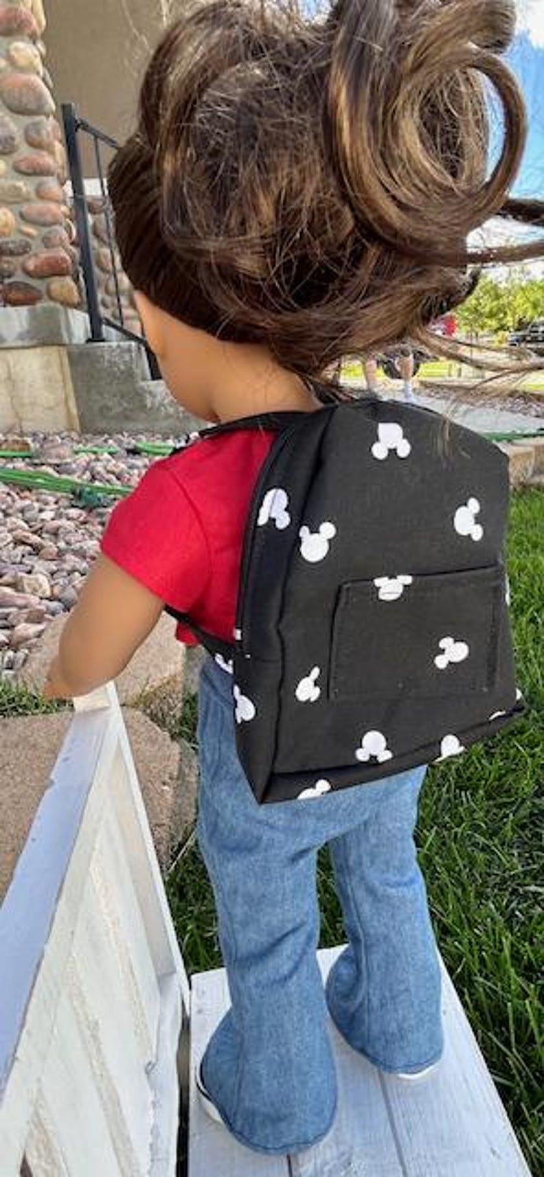 May include: A black backpack with white Mickey Mouse heads printed all over it. The backpack has a small pocket on the front. The backpack is being worn by a doll with brown hair and a red shirt.