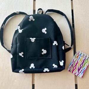 May include: A small black backpack with white Mickey Mouse silhouettes all over it. The backpack has adjustable straps and a zipper closure.