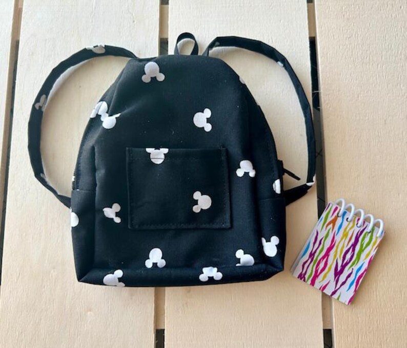 May include: A small black backpack with white Mickey Mouse silhouettes all over it. The backpack has adjustable straps and a zipper closure.