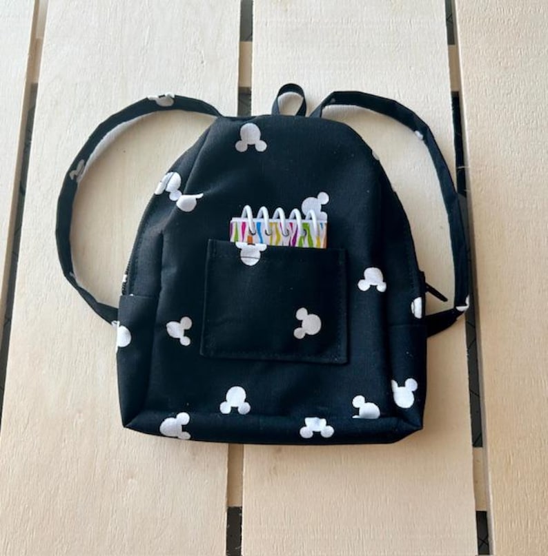 May include: A small black backpack with white Mickey Mouse ears printed all over. The backpack has a zipper closure and a front pocket with a small notebook inside.