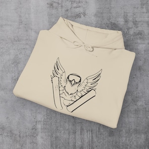 May include: A cream-colored hoodie featuring a black graphic of an owl wearing goggles and skis with outstretched wings. The hoodie has a drawstring hood and is folded on a gray surface.