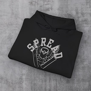 May include: Black hoodie with a white graphic design. The design features the word "SPREAD" above an owl wearing goggles, with skis on either side. The hoodie is folded on a gray surface.