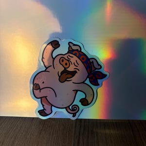 Puede incluir: Una pegatina con un cerdo de dibujos animados con un pañuelo rojo y azul, con la lengua fuera y los brazos levantados. El cerdo es de color rosa claro con contornos negros. El fondo es un material holográfico iridiscente.