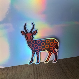 Puede incluir: Una pegatina con un ciervo estilizado con cara y cuernos rojos. El cuerpo del ciervo está cubierto de círculos naranjas y azules. La pegatina tiene un borde blanco y está sobre un fondo holográfico.