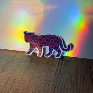 May include: A sticker featuring a stylized leopard design. The leopard has a blue body with red circles and red details. The sticker is on a wooden surface with a holographic background.