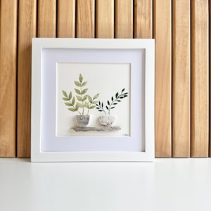 May include: A framed watercolor painting featuring two potted plants with green leaves. The plants are in decorative pots, one with a blue and white pattern, and the other with a speckled design. The artwork is set against a light background.